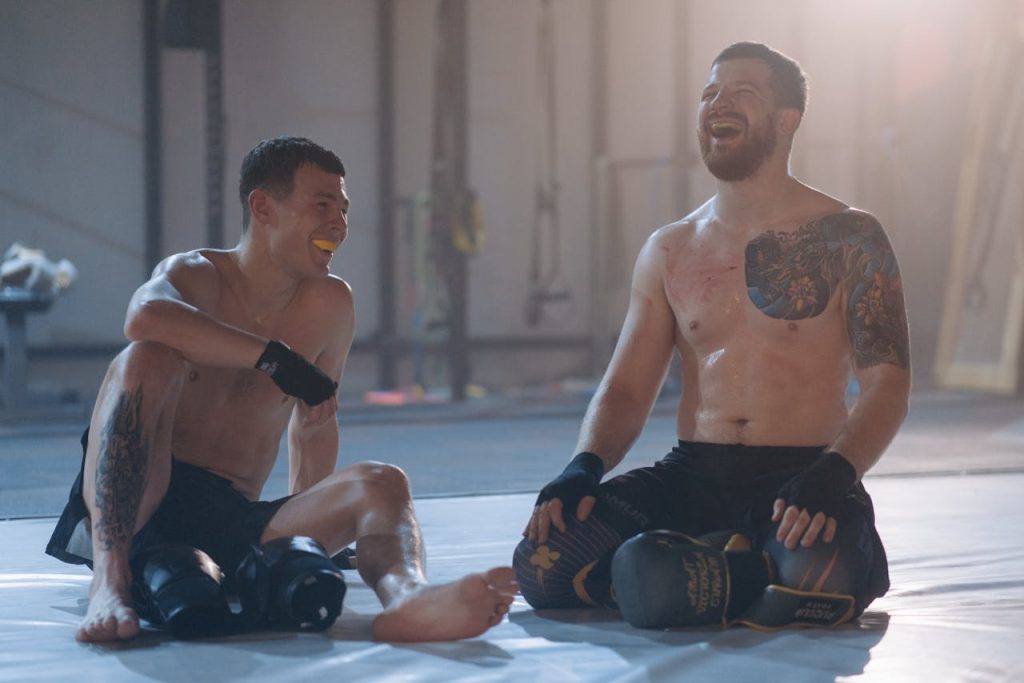 Two shirtless men with tattoos and mouthguards share a laugh in a gym setting, showcasing camaraderie.